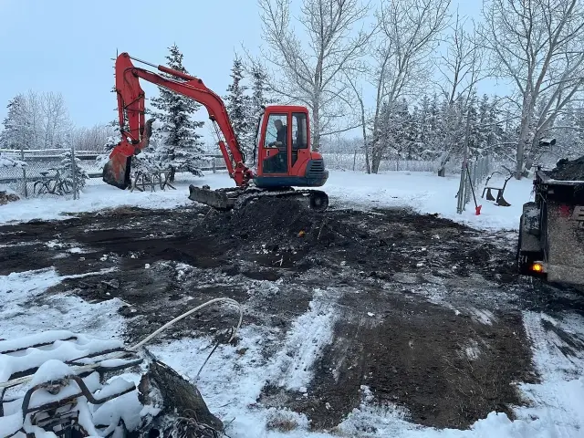 Building contractors Calgary use an excavator to dig into snowy ground on a cloudy day