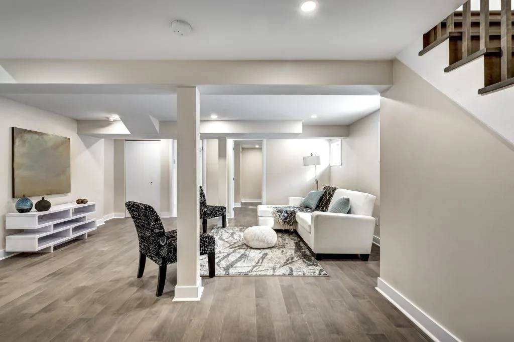 Basement renovation featuring a bright living area with a white sofa, patterned armchairs, and a large rug on light wood flooring